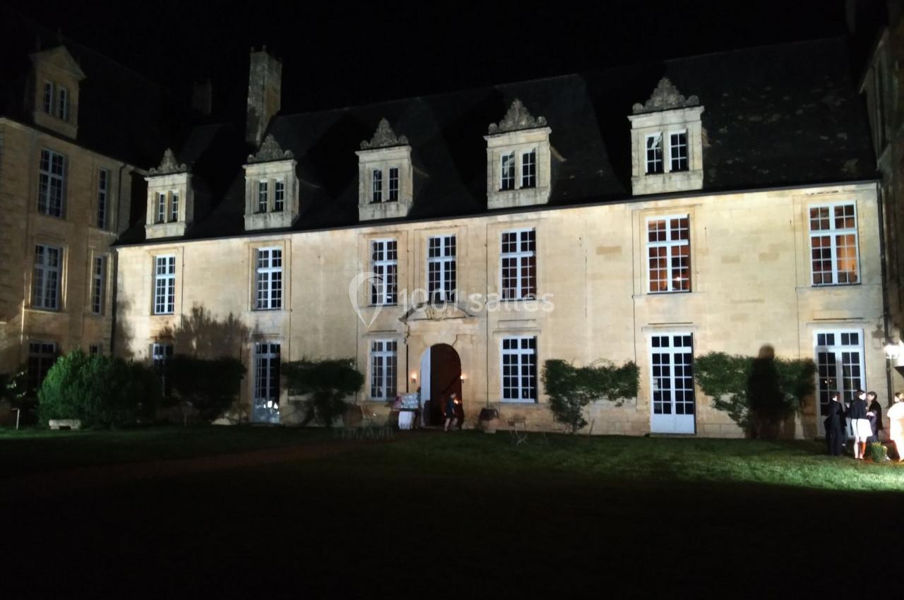 Façade d'un bâtiment historique en pierre éclairée la nuit, entourée de végétation et d'une pelouse.