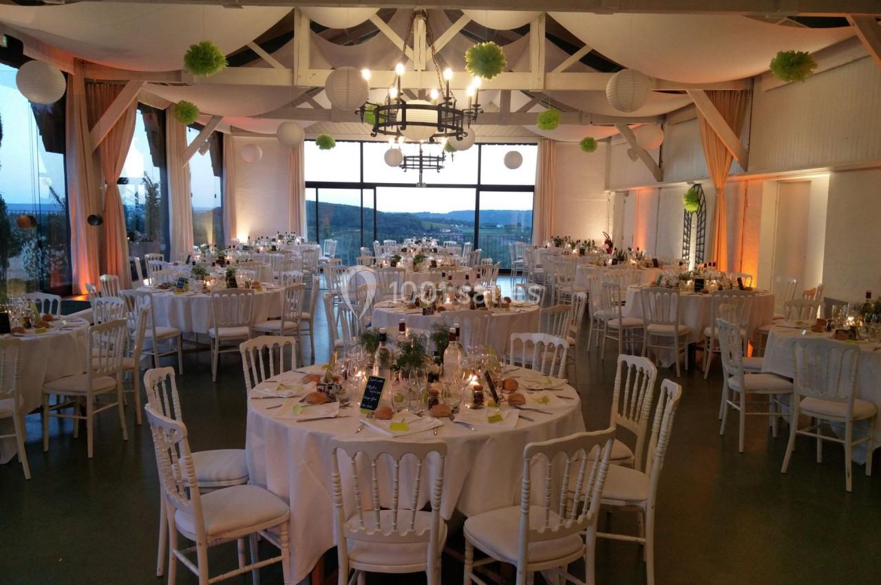 Salle de réception décorée pour un événement, avec tables rondes dressées, chandeliers et vue sur l'extérieur.