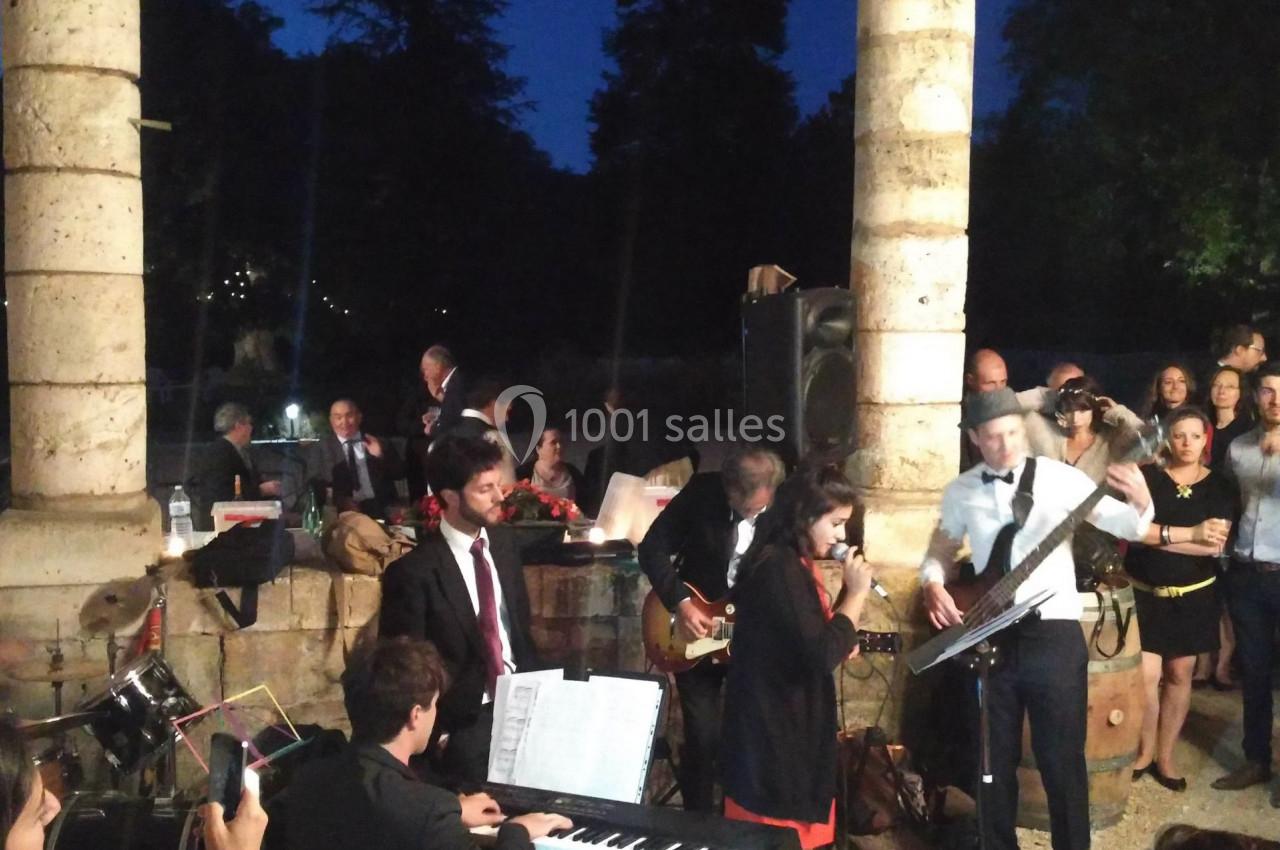 Un groupe de musiciens joue en plein air devant un public, avec une chanteuse au micro et un pianiste au premier plan.