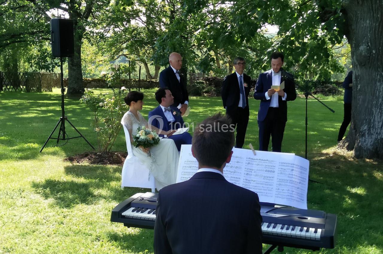 Un mariage en extérieur avec un couple assis, un pianiste jouant et un homme lisant un discours devant des invités.