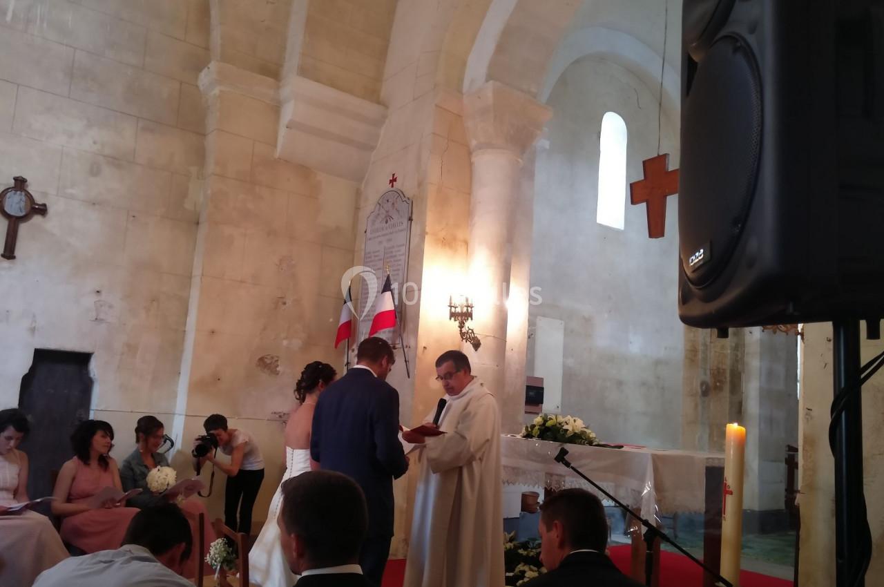 Un couple échange ses vœux devant un prêtre dans une église, entouré d'invités assis et de décorations simples.