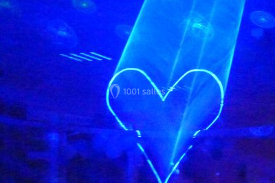 Façade en pierre éclairée par des lumières violettes et bleues, avec des tables en métal noir au premier plan.