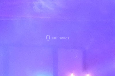 Façade en pierre éclairée par des lumières violettes et bleues, avec des tables en métal noir au premier plan.