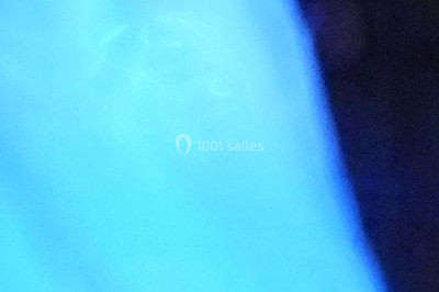 Façade en pierre éclairée par des lumières violettes et bleues, avec des tables en métal noir au premier plan.