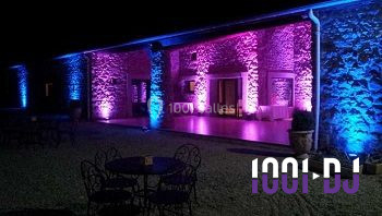 Façade en pierre éclairée par des lumières violettes et bleues, avec des tables en métal noir au premier plan.