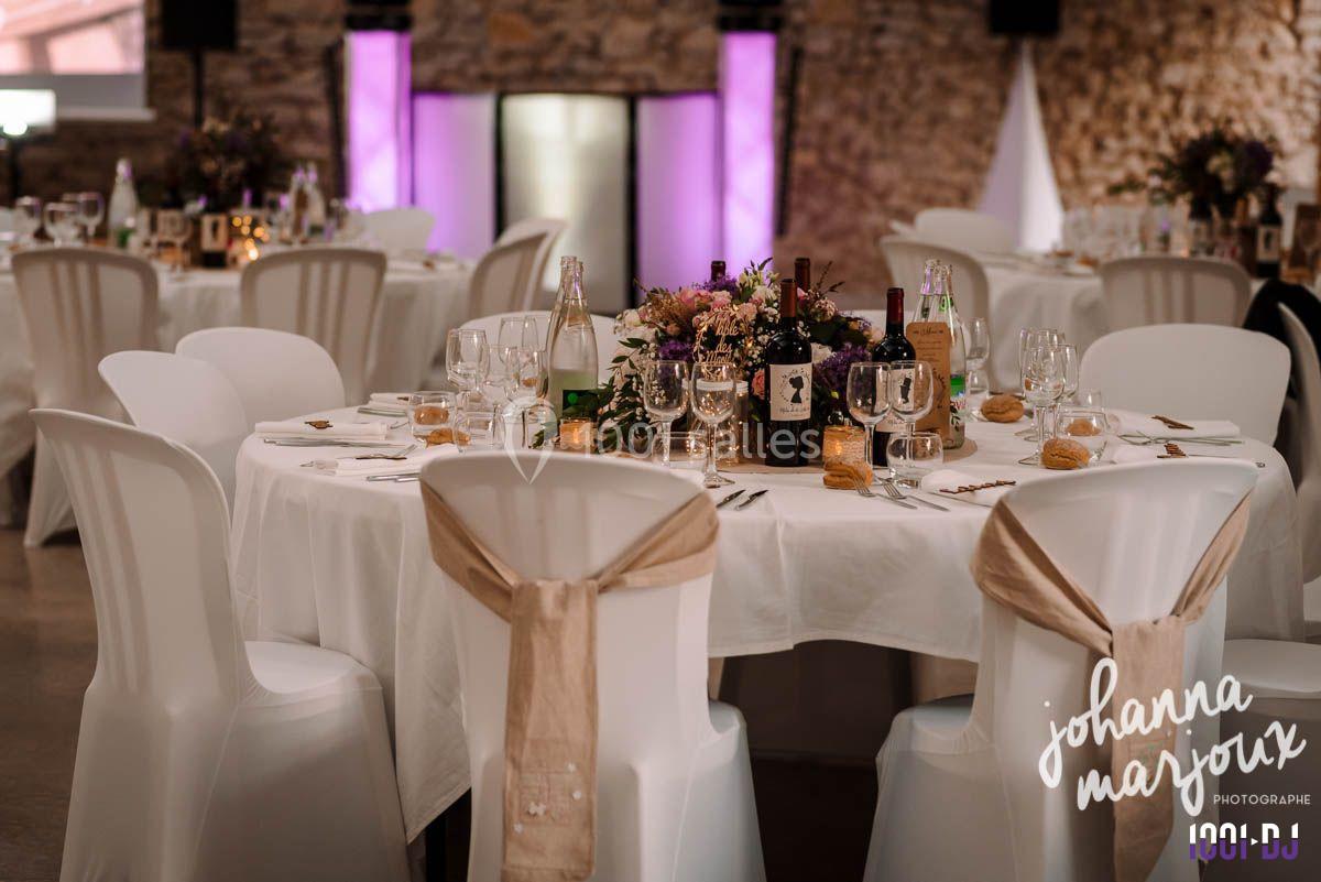 Salle de réception décorée avec des tables rondes, nappes blanches, chaises ornées de rubans et centre de table fleuri.