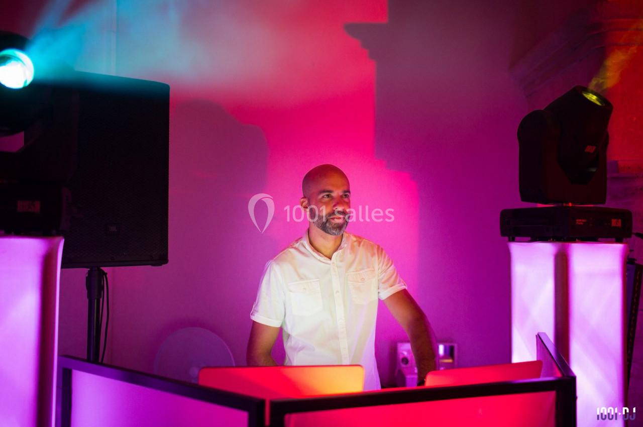Un DJ en chemise blanche mixe de la musique derrière une table éclairée par des lumières colorées.