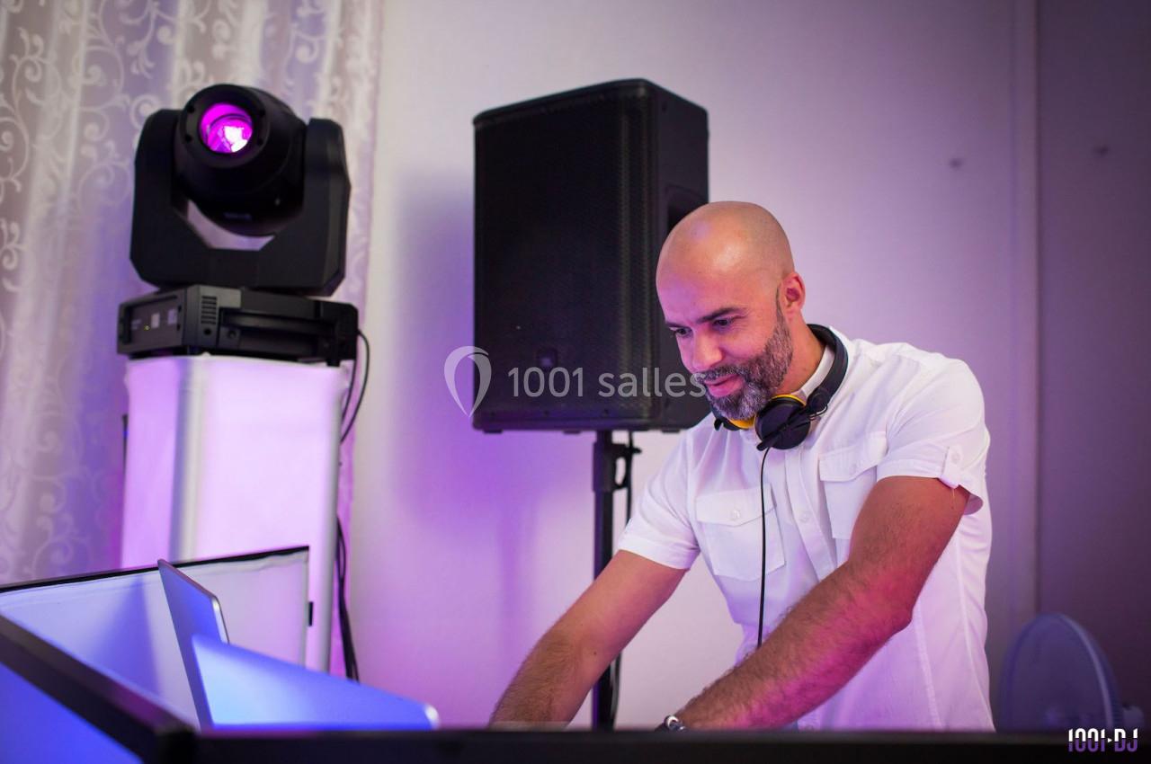 Un DJ en chemise blanche mixe de la musique sur une table de mixage, éclairé par des lumières violettes.