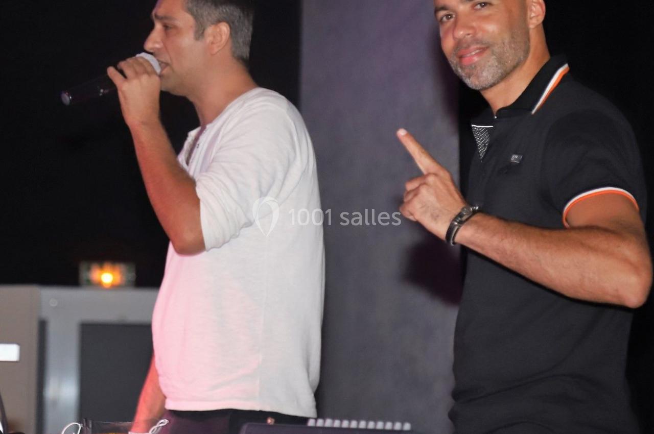 Deux hommes sur scène, l'un chante au micro tandis que l'autre pointe du doigt près d'un équipement audio.