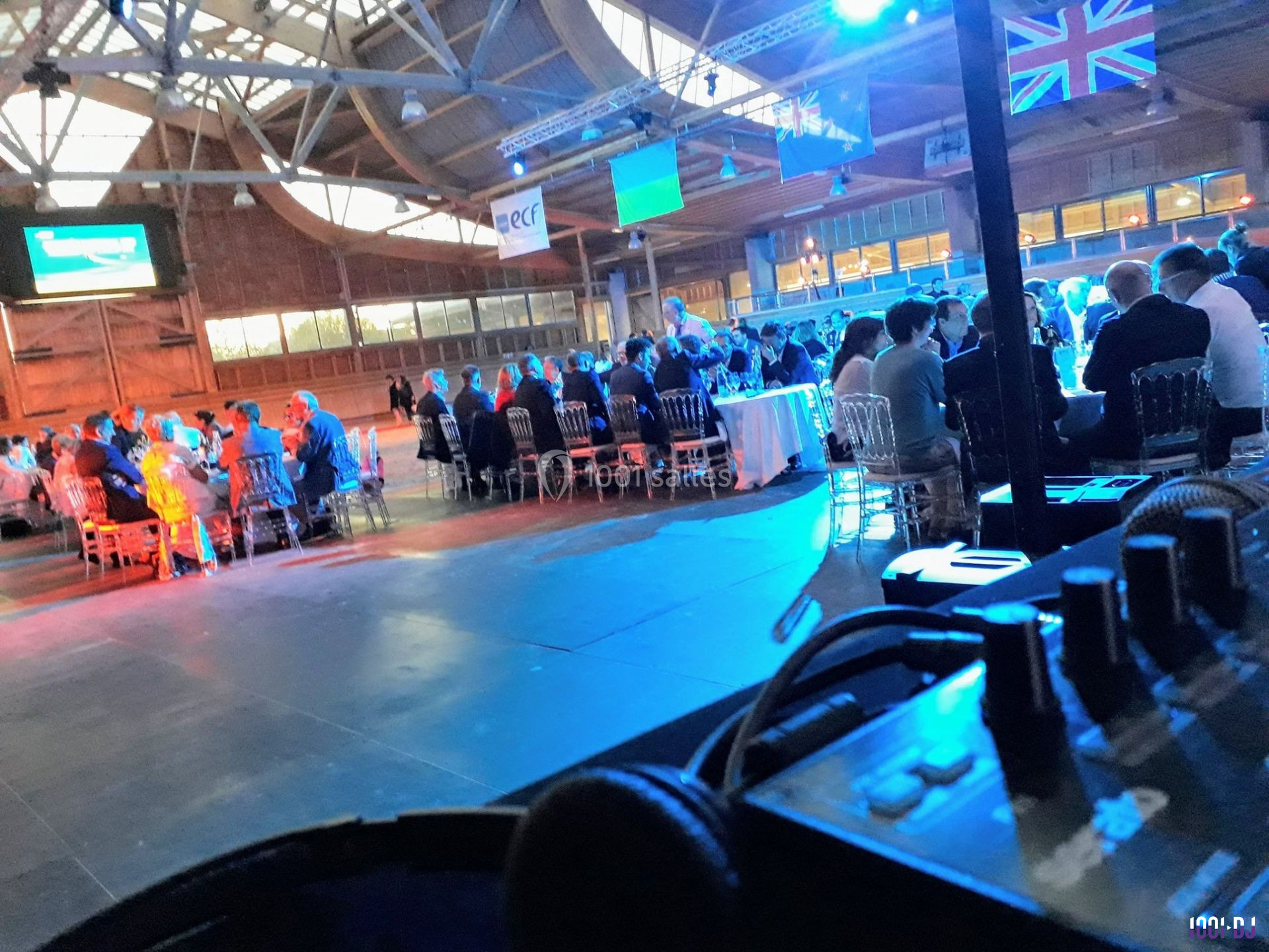 Salle de conférence avec des tables rondes occupées par des participants, éclairage bleu et drapeaux suspendus.