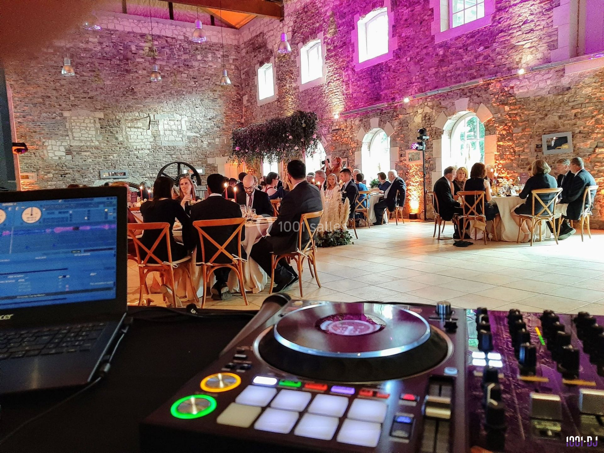 Salle de réception en pierre avec invités assis à des tables rondes, vue depuis une table de mixage de DJ au premier plan.