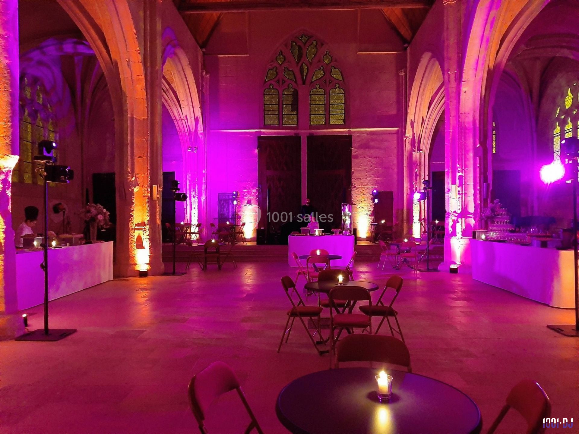 Salle voûtée éclairée de lumières violettes, avec tables et chaises disposées pour un événement.