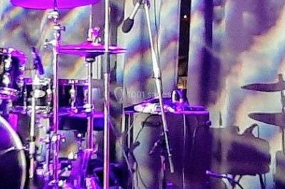 Scène de concert avec batterie, guitares, claviers et micros sous un éclairage coloré.