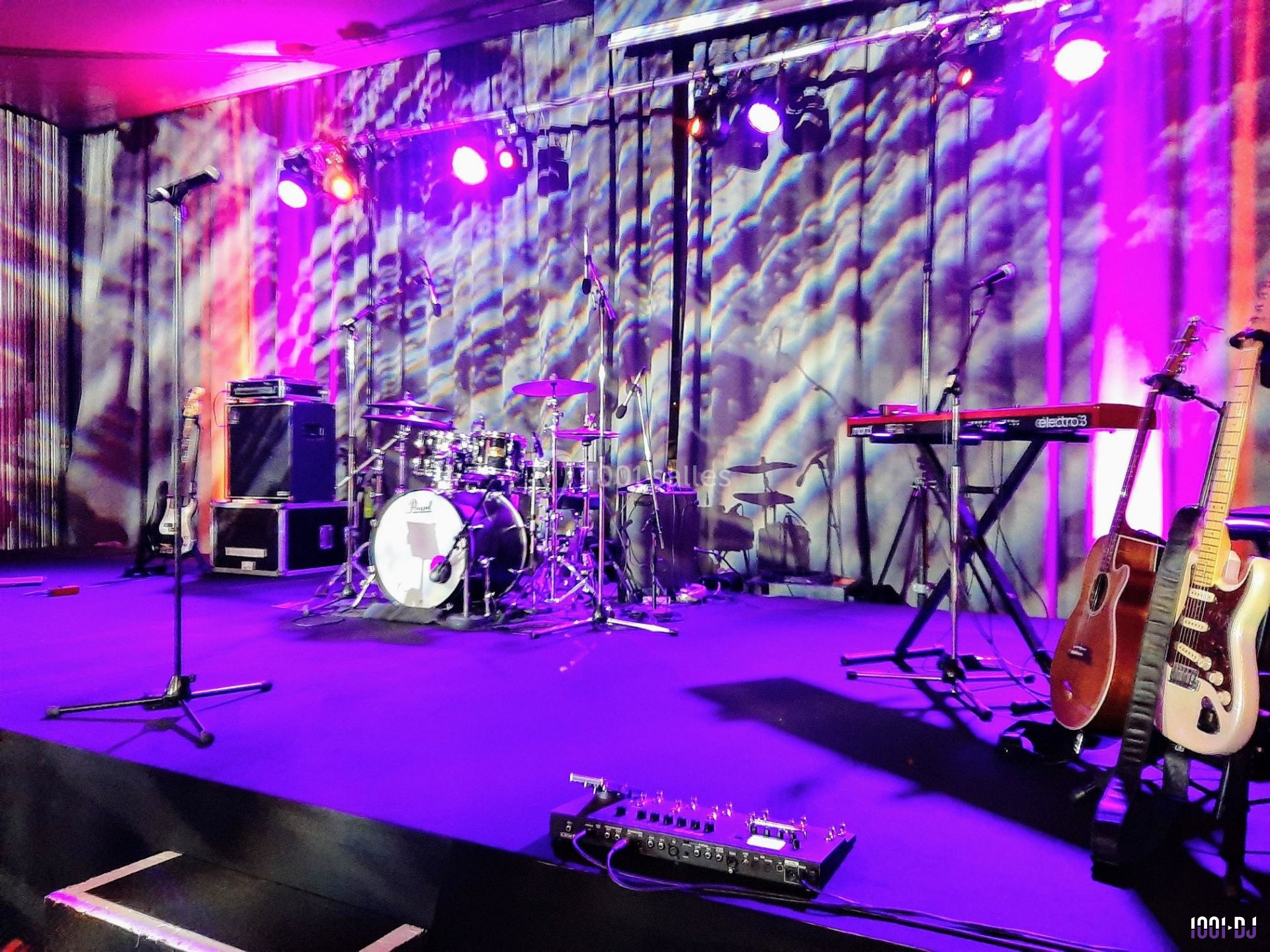 Scène de concert avec batterie, guitares, claviers et micros sous un éclairage coloré.