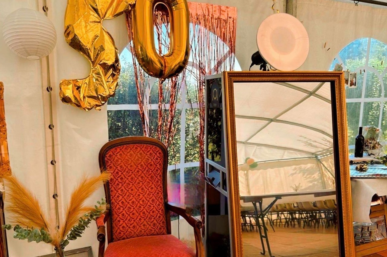 Fauteuil rouge, miroir doré, ballons ’50’ et décor festif dans une tente avec vue sur un jardin.