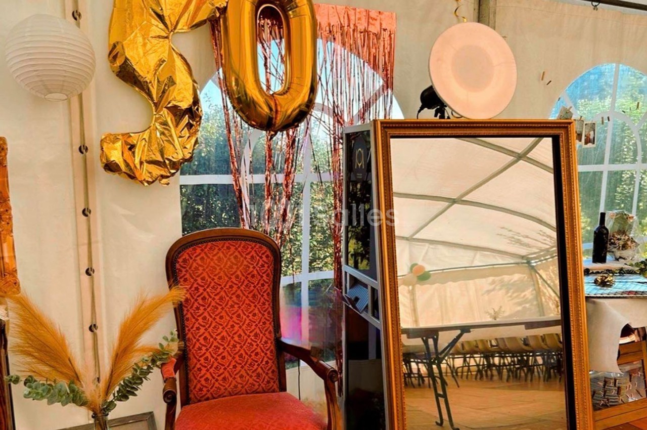 Fauteuil rouge, miroir doré, ballons ’50’ et décor festif dans une tente avec vue sur un jardin.