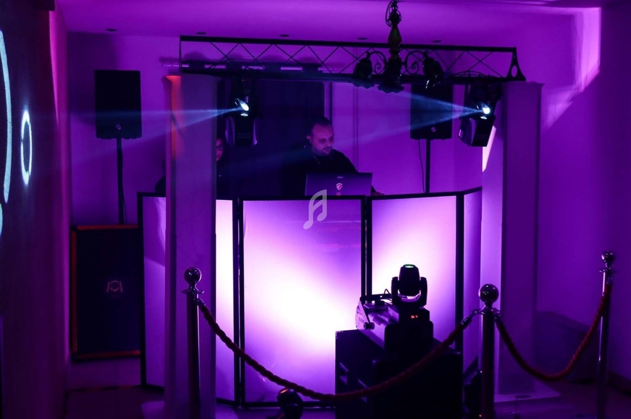 Un DJ mixe derrière une table éclairée de lumières violettes, entourée d'équipements audio et de projecteurs.