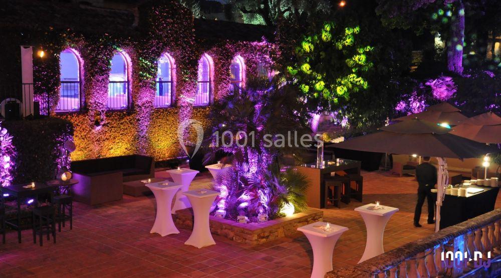 Cour intérieure éclairée avec des lumières colorées, tables hautes décorées et végétation luxuriante en soirée.