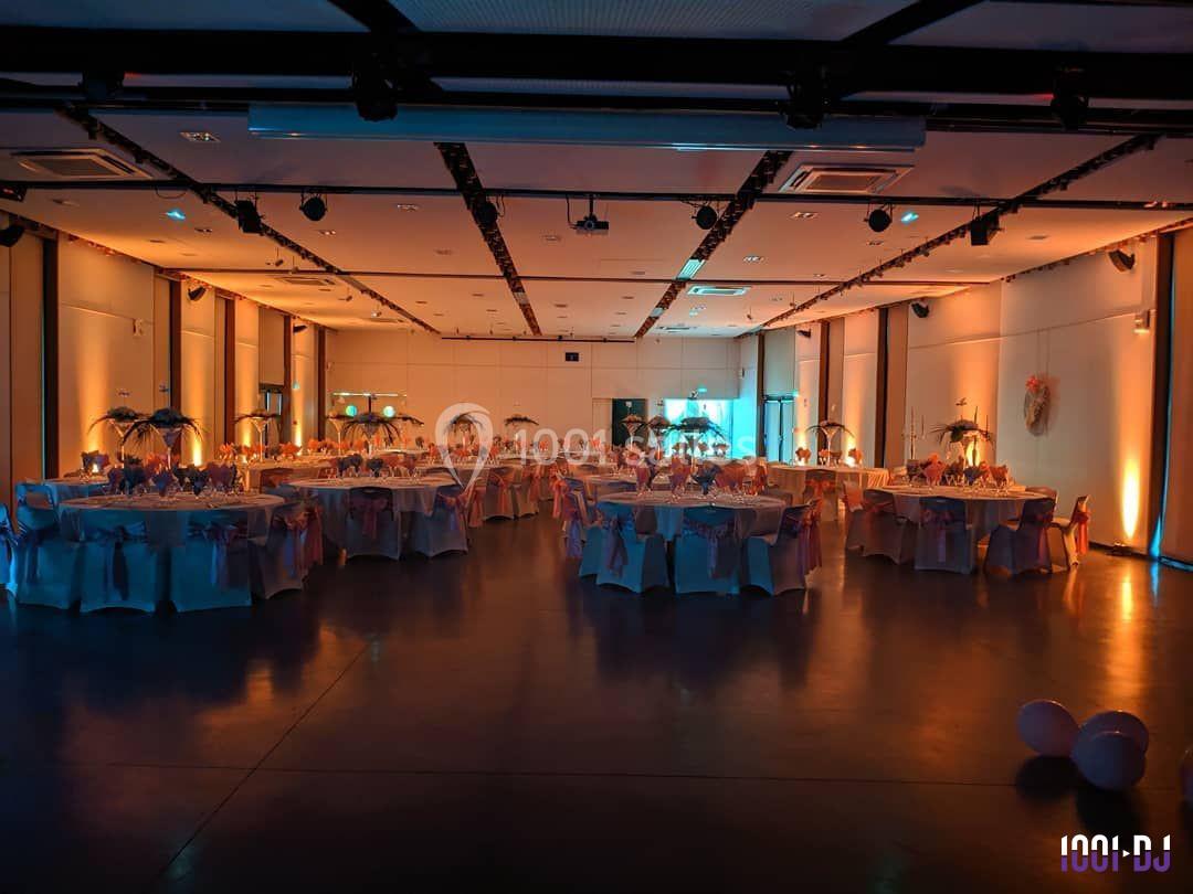 Salle de réception décorée avec des tables rondes, nappes blanches et éclairage tamisé orangé.