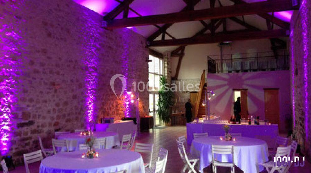 Salle de réception en pierre avec éclairage violet, tables rondes décorées et chaises blanches.