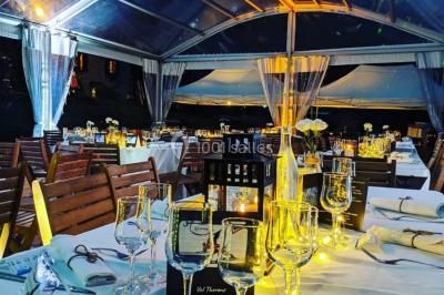 Salle de réception en plein air sous une verrière, avec tables décorées, nappes blanches et éclairage chaleureux.