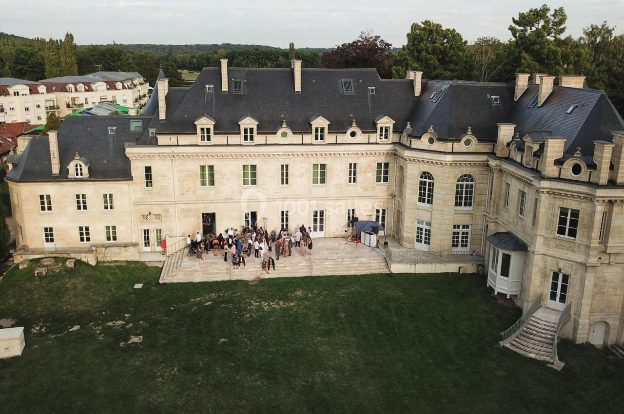 Vue aérienne d'un bâtiment historique en pierre avec des invités rassemblés sur une terrasse, entouré de verdure.