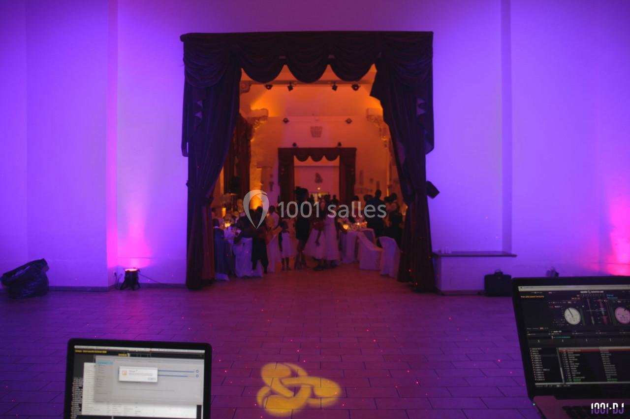 Salle de réception éclairée en violet et orange, avec des invités rassemblés au fond et des équipements de DJ au premier…