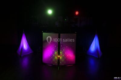 Salle de réception éclairée avec des lumières colorées, vue depuis une table de DJ équipée d'un ordinateur et de platines.