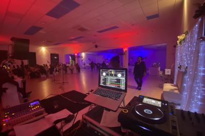 Salle de réception éclairée avec des lumières colorées, vue depuis une table de DJ équipée d'un ordinateur et de platines.