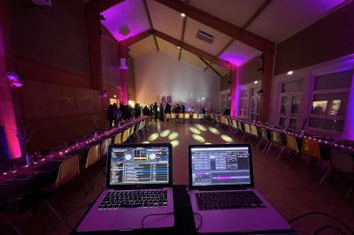 Salle de réception éclairée avec des lumières colorées, vue depuis une table de DJ équipée d'un ordinateur et de platines.