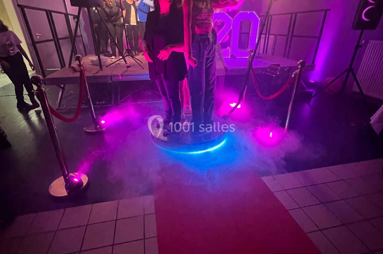 Deux personnes posent sur une plateforme lumineuse entourée de fumée, avec une scène décorée en arrière-plan.