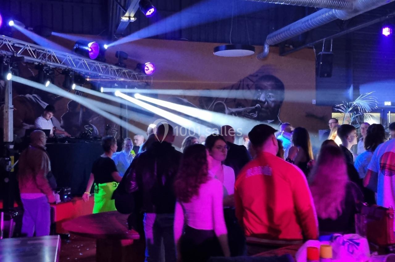 Des personnes dans une salle animée avec un DJ sur scène, éclairée par des projecteurs colorés.