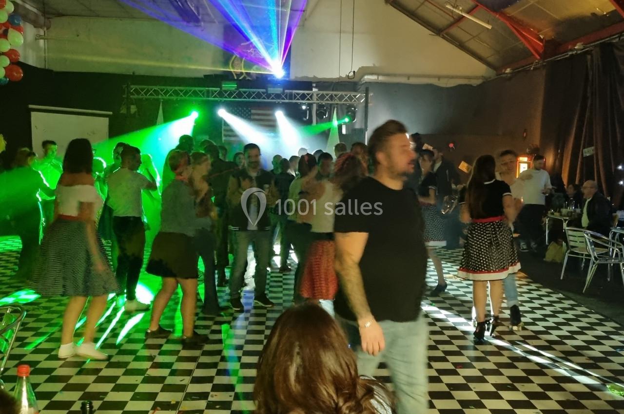 Des personnes dansent sur une piste à damier éclairée par des lumières colorées dans une ambiance festive en intérieur.