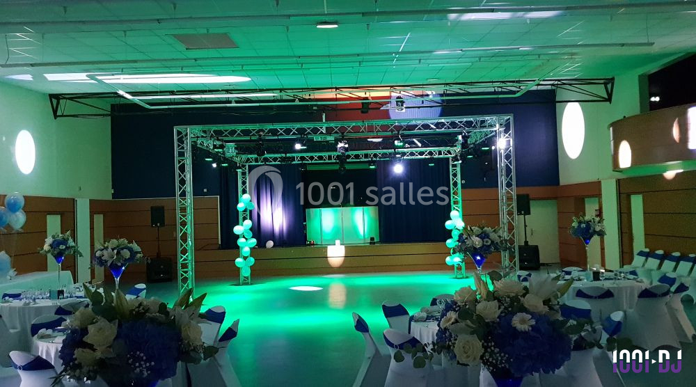 Salle de réception décorée avec des tables dressées, des fleurs et une scène éclairée par des projecteurs colorés.