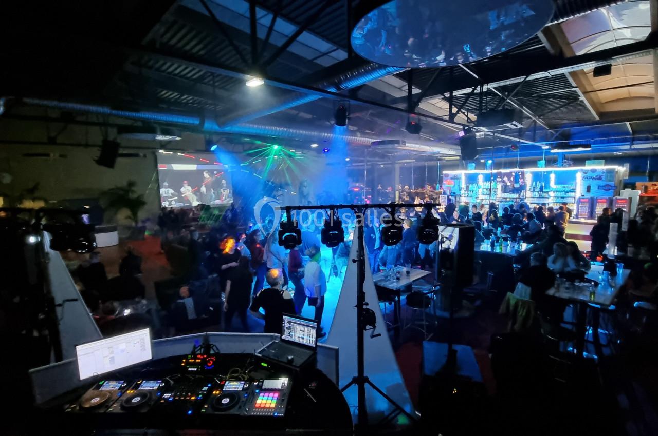 Salle de fête animée avec des jeux de lumière, une piste de danse, un DJ et des invités assis ou debout.