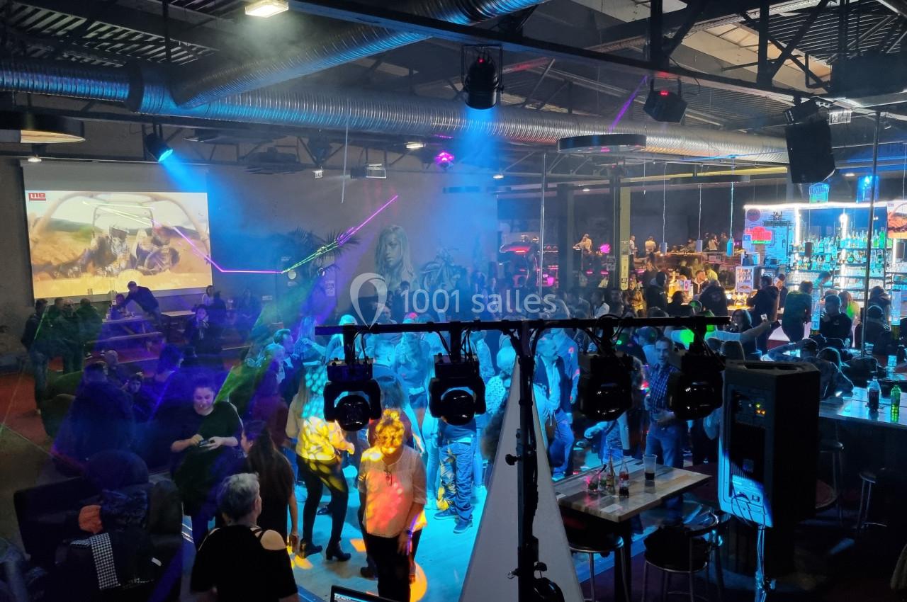 Personnes dansant et discutant dans un bar animé avec éclairages colorés et un écran projetant une vidéo.