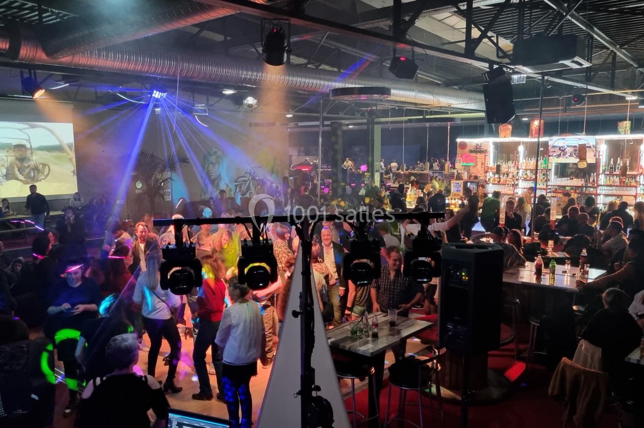 Salle animée avec des personnes dansant et discutant, éclairage coloré, bar illuminé en arrière-plan.