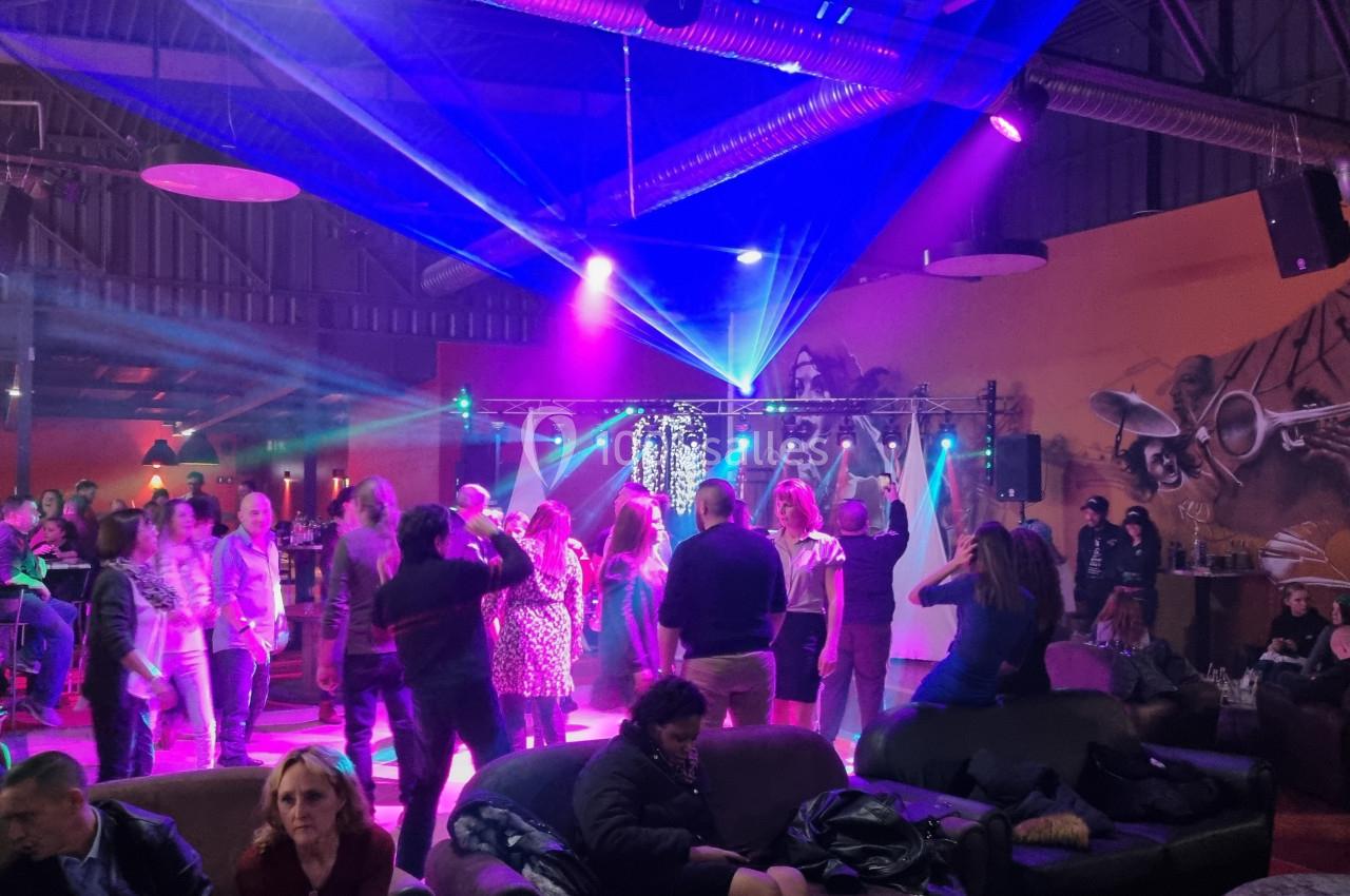 Des personnes dansent sous des jeux de lumière colorés dans une salle avec un décor mural musical et des spectateurs assis.