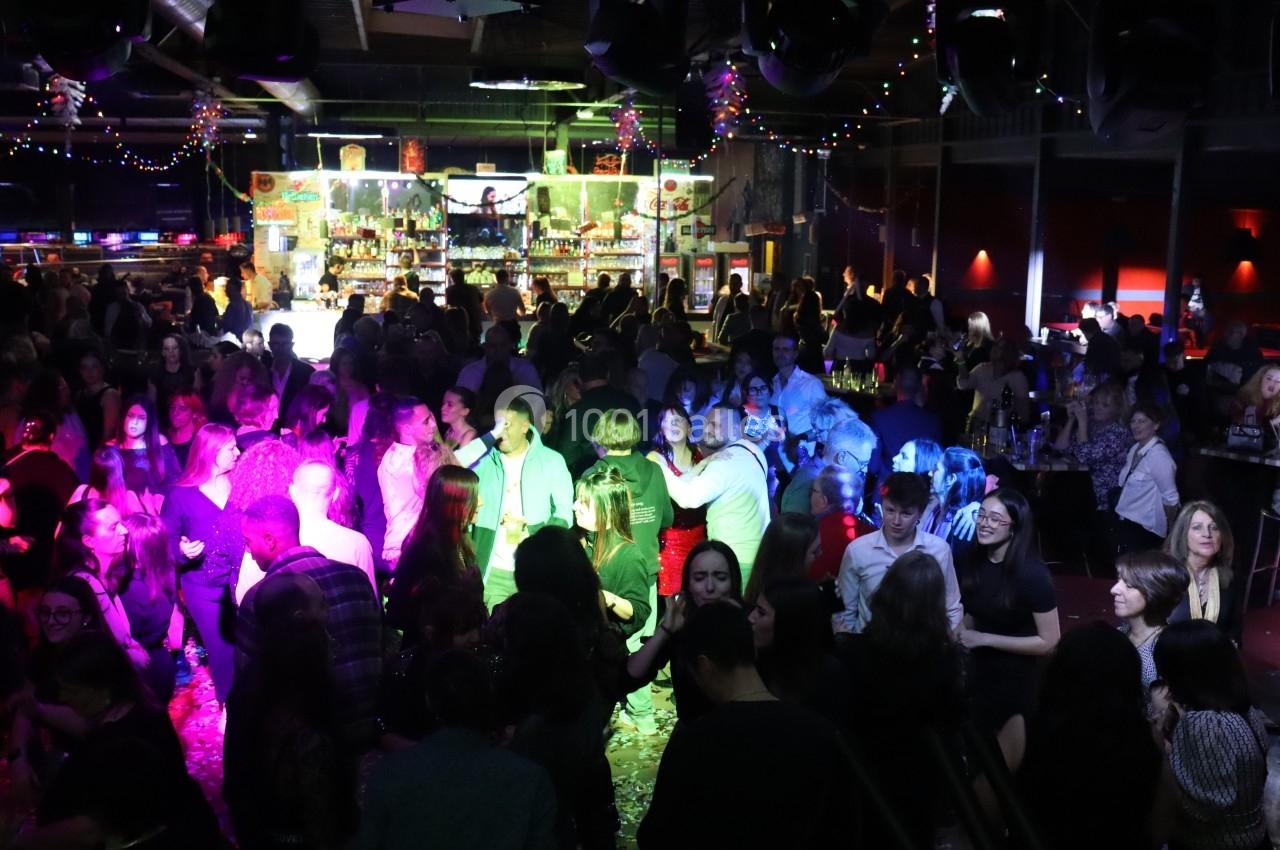 Salle de fête animée avec des personnes dansant sous des lumières colorées, un bar lumineux en arrière-plan.