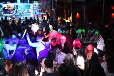 Vue depuis une table de mixage sur une salle éclairée par des lumières colorées, avec des invités assis et debout.