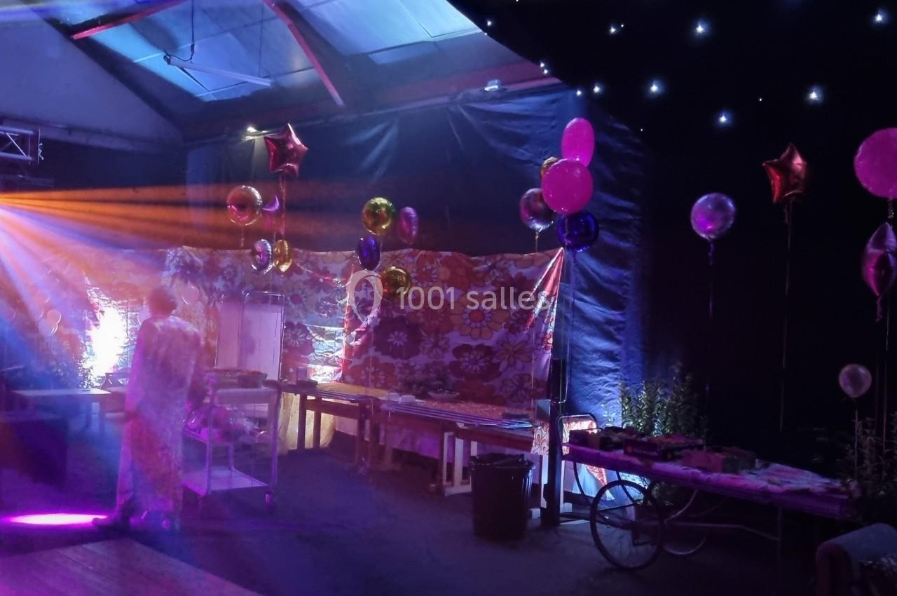 Salle décorée pour une fête avec ballons colorés, lumières tamisées et effets lumineux sur une scène.