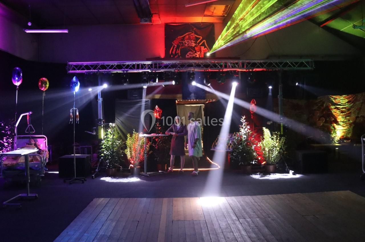 Salle décorée avec des plantes, lumières colorées et lasers, avec deux personnes au centre sous un projecteur.
