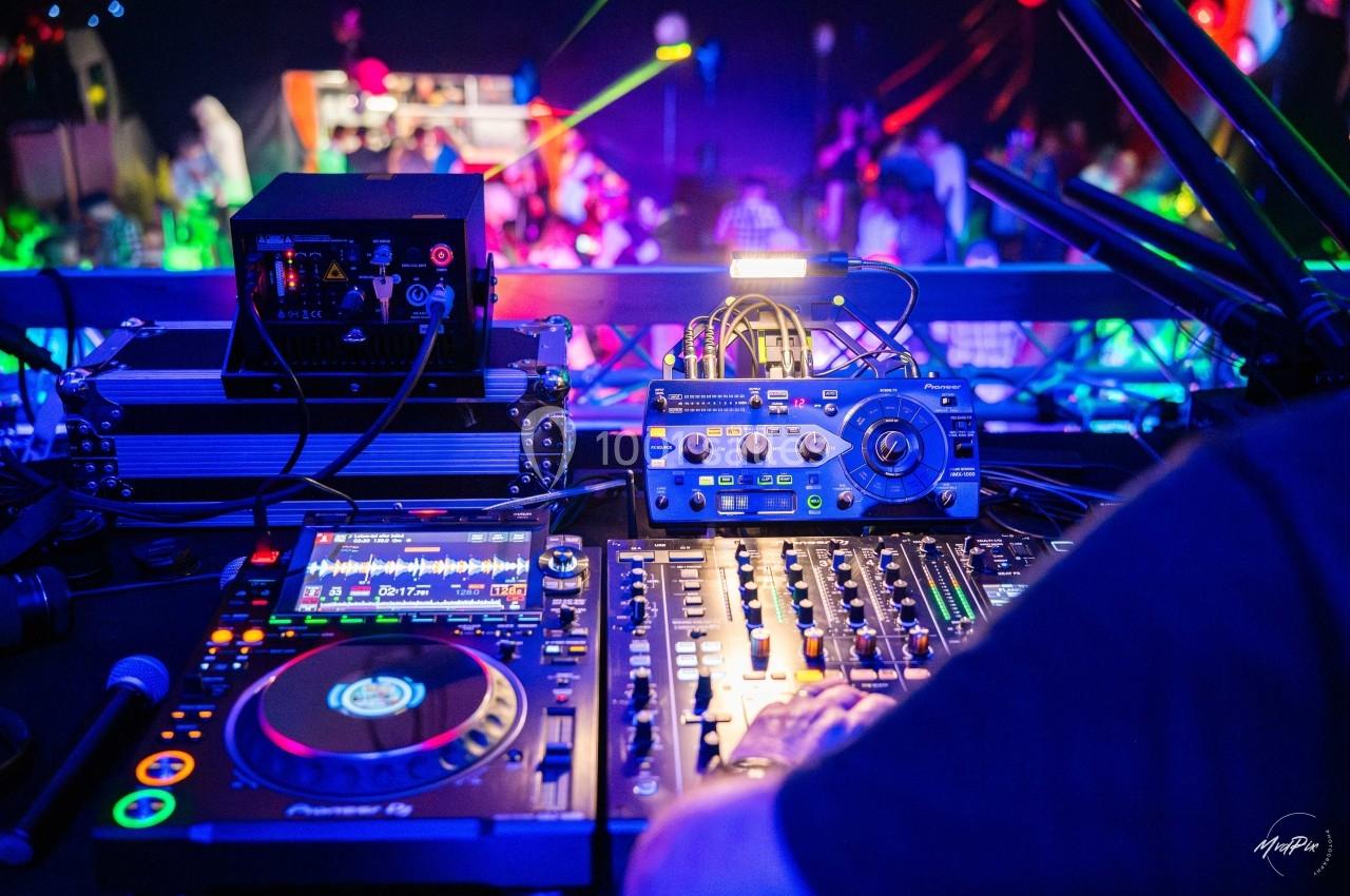 Vue rapprochée d'une table de mixage et d'équipements DJ, avec une piste de danse éclairée en arrière-plan.