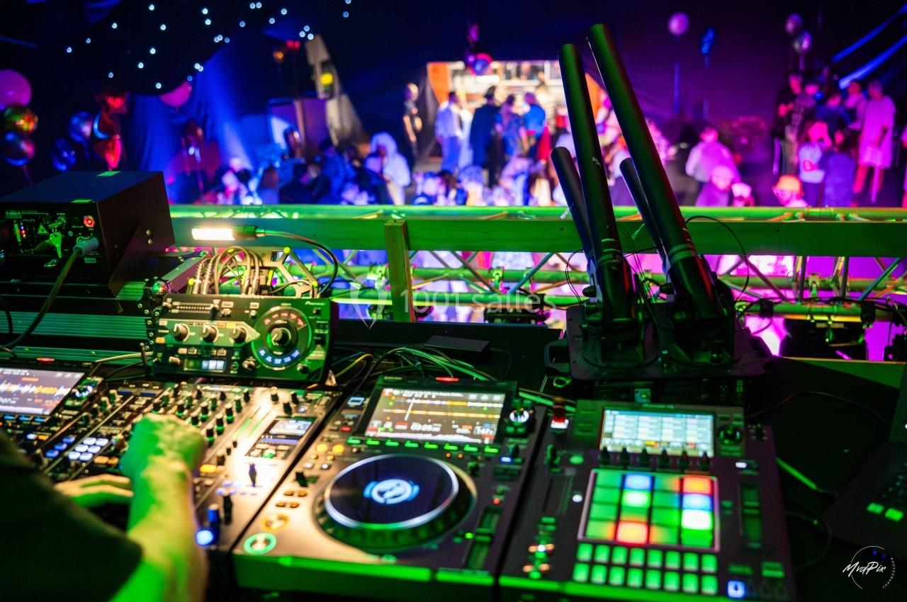 Vue depuis une cabine de DJ avec des platines et des lumières colorées, face à une foule dansante en arrière-plan.