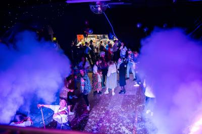 Vue depuis une table de mixage sur une salle éclairée par des lumières colorées, avec des invités assis et debout.