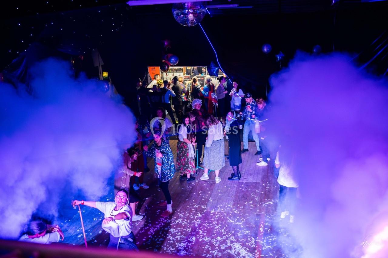 Personnes dansant et socialisant dans une salle festive éclairée par des lumières colorées, avec de la fumée et des…