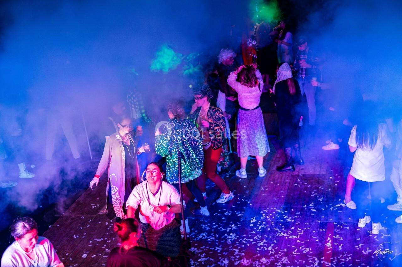 Des personnes dansent sous des lumières colorées et des effets de fumée dans une ambiance festive.