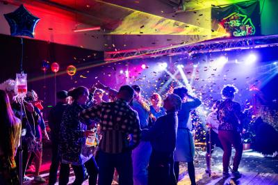 Vue depuis une table de mixage sur une salle éclairée par des lumières colorées, avec des invités assis et debout.