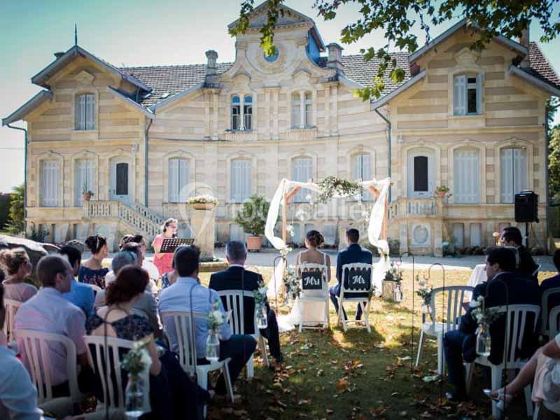 Cérémonie de mariage en plein air devant une grande maison ancienne, avec invités assis et arche décorée.