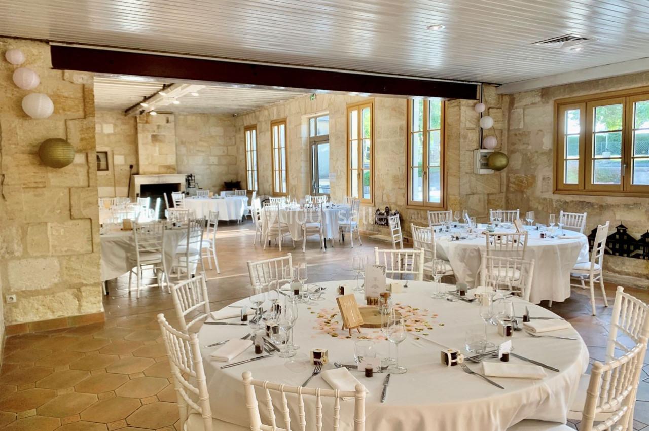 Salle de réception lumineuse avec murs en pierre, tables rondes dressées et chaises blanches.